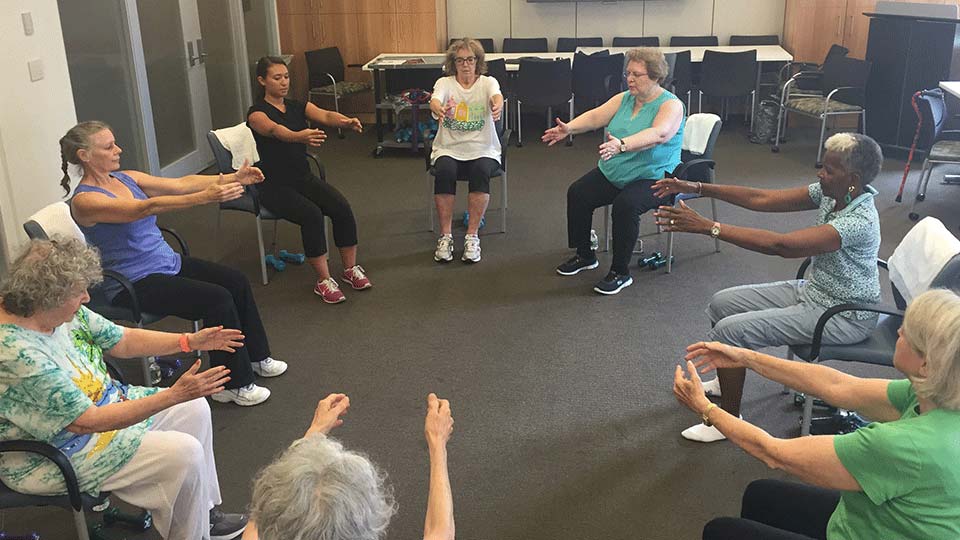 Group of people in large room sitting in chairs in a circle performing an abdominal activation exercise