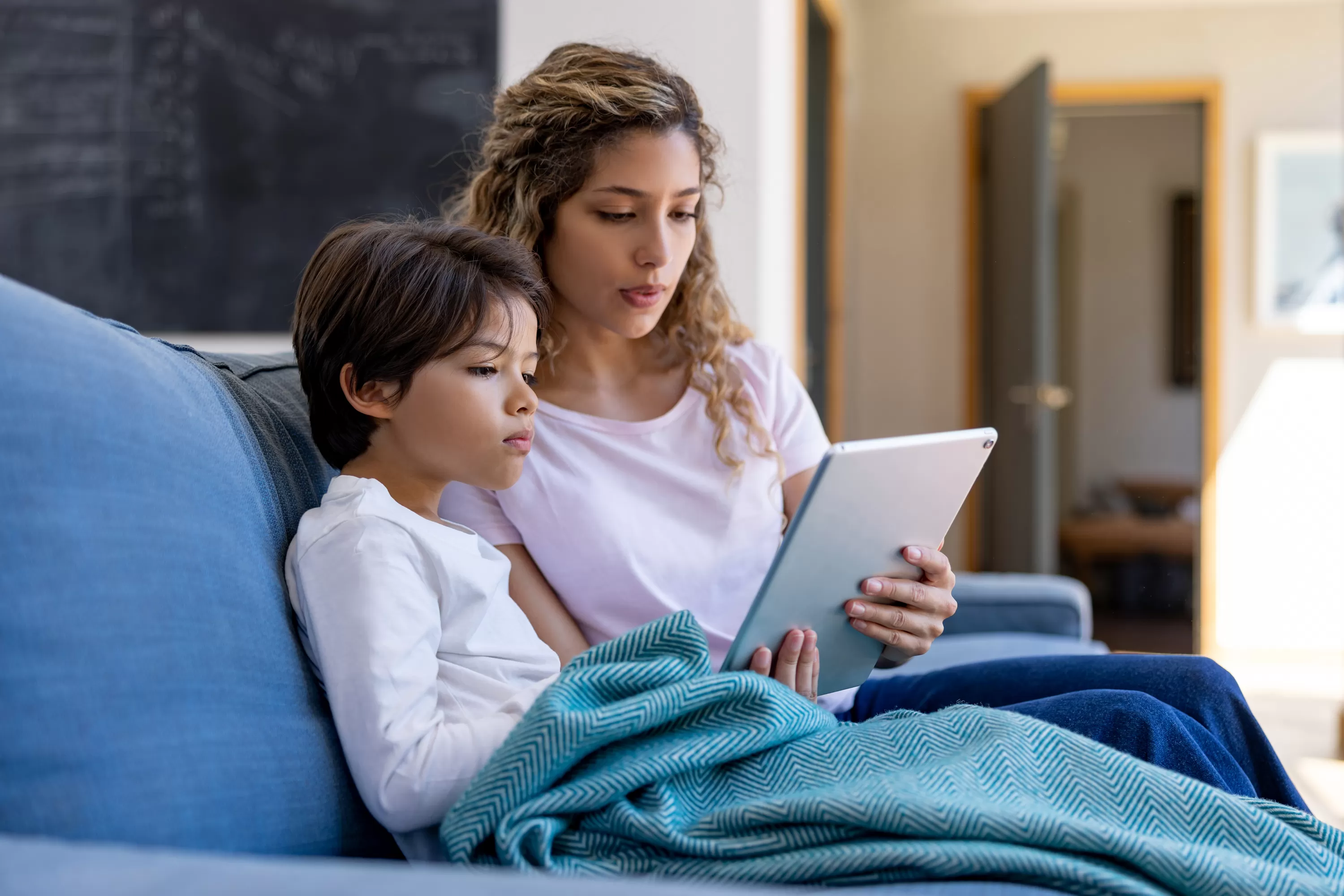 Child wrapped in blanket seated on couch next to mother holding a tablet device for both of them to view
