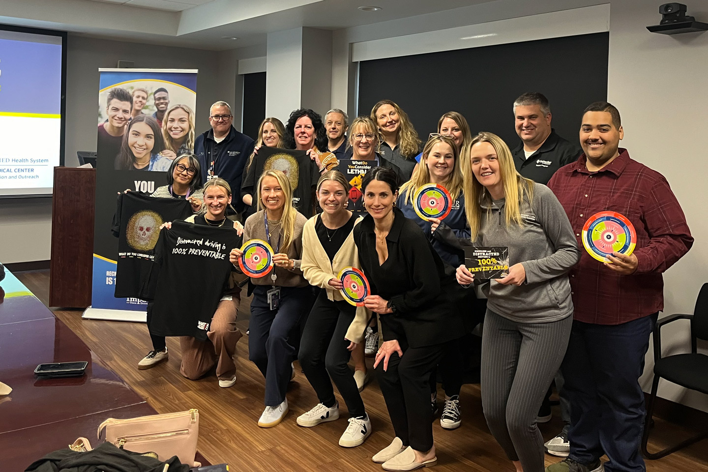 A group of people pose in a classroom, holding signs and shirts with safety messages about preventing distracted driving.