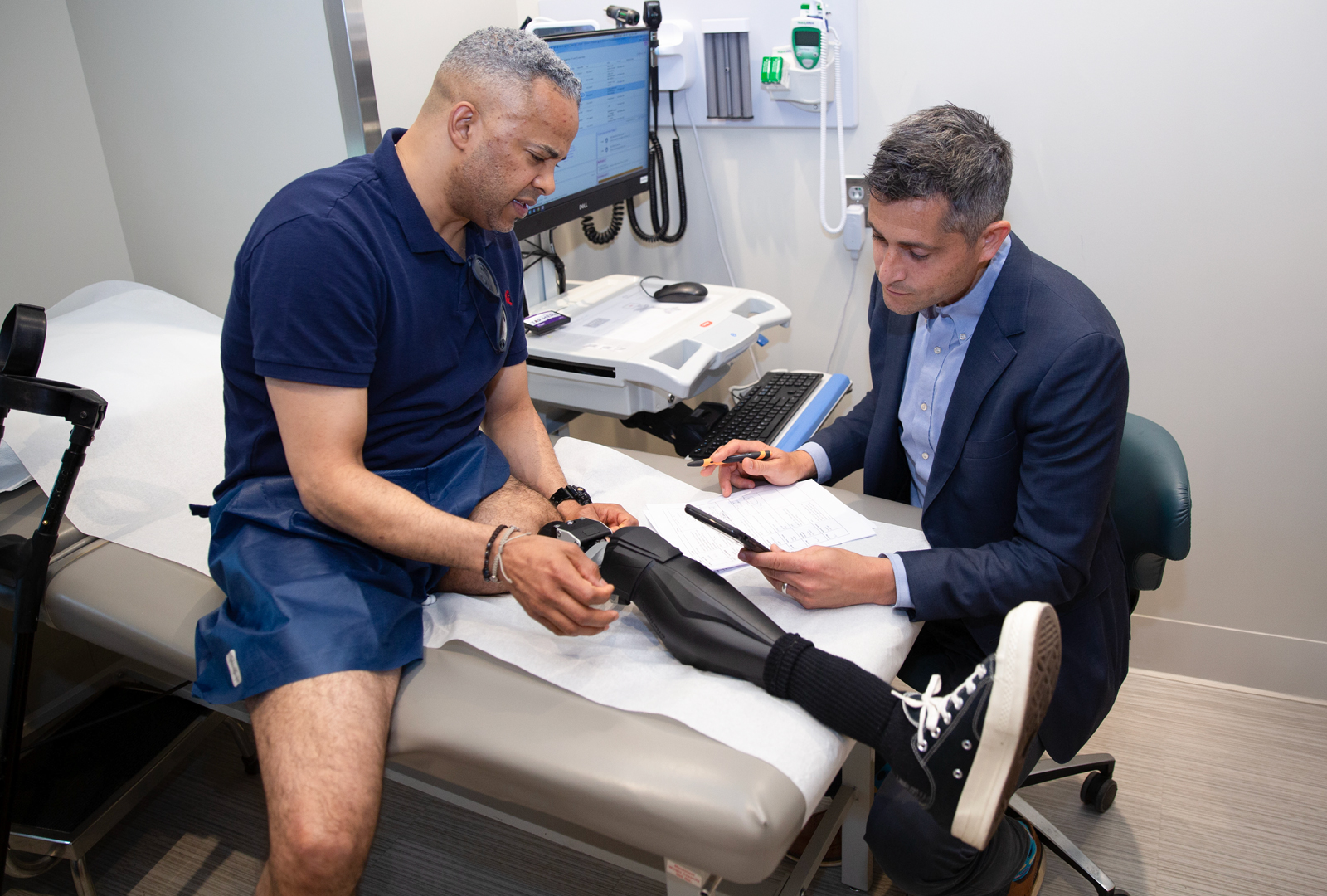 A male patient, left, sits on an exam bed while a male doctor, right, examines his prosthesis.