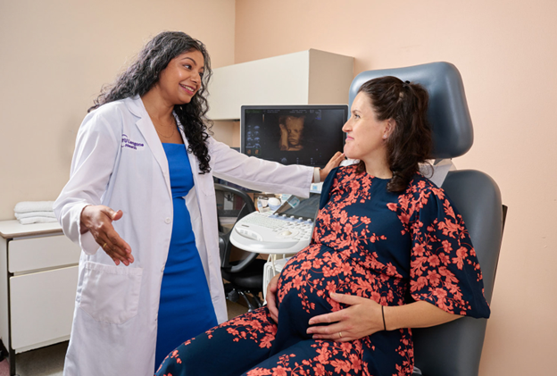 A healthcare provider in a white coat talks with a visibly pregnant patient seated in an exam chair beside an ultrasound machine displaying a fetal image.