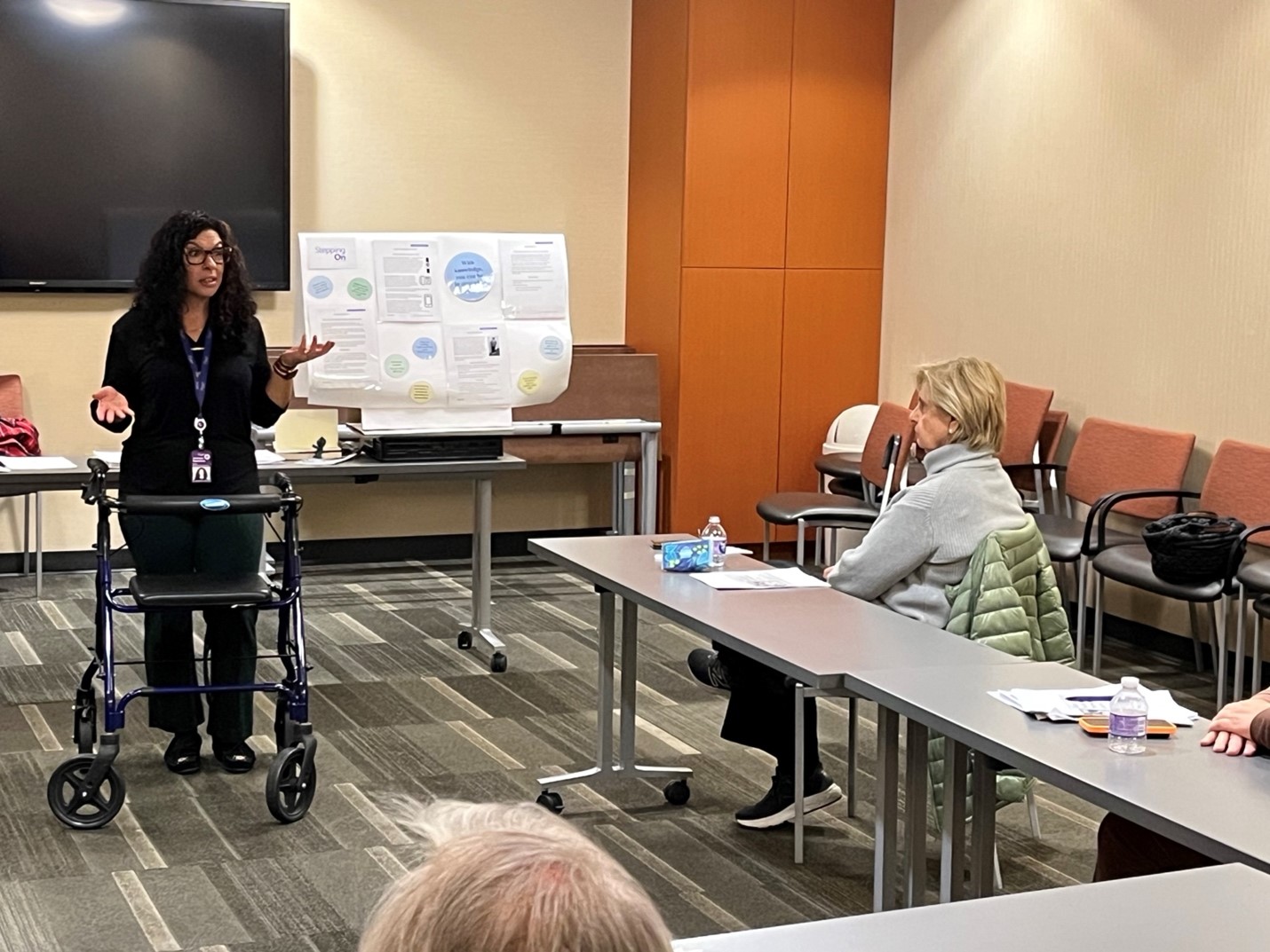 Dr. Jeannine Nonaillada stands at the front of a classroom with a walker while speaking to several people seated at tables.