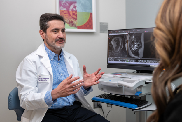 Doctor Jose Peiro, in a white coat, gestures while speaking with a patient in an exam room, seated beside a computer monitor displaying medical scan images.