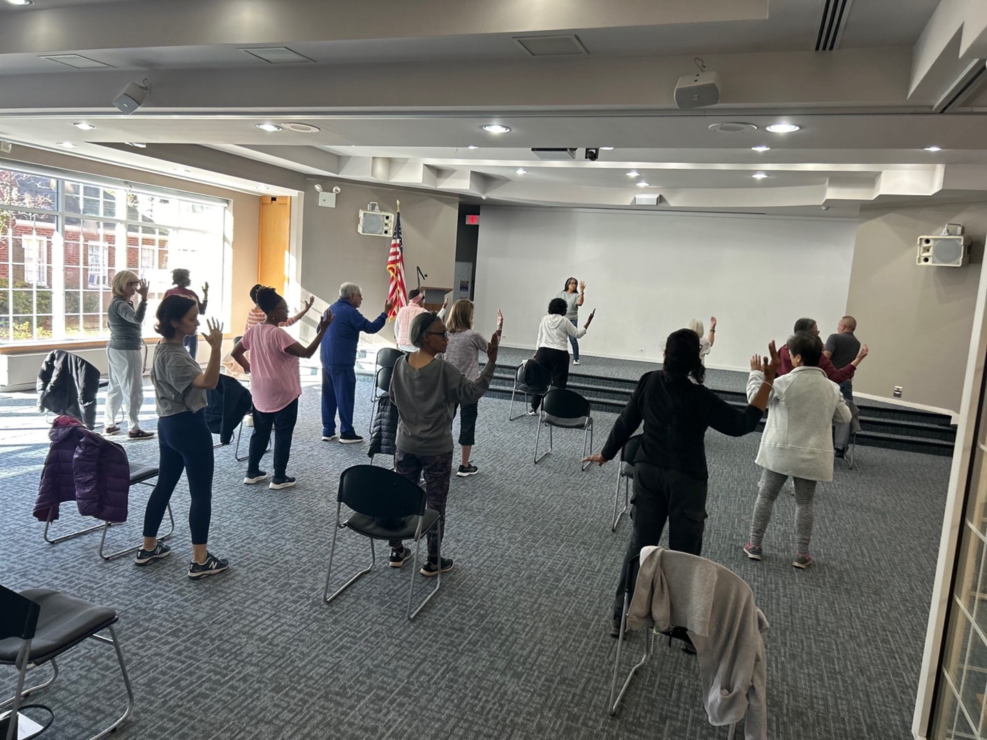 A woman teaches tai chi to a group of people, with each participant standing by a chair.