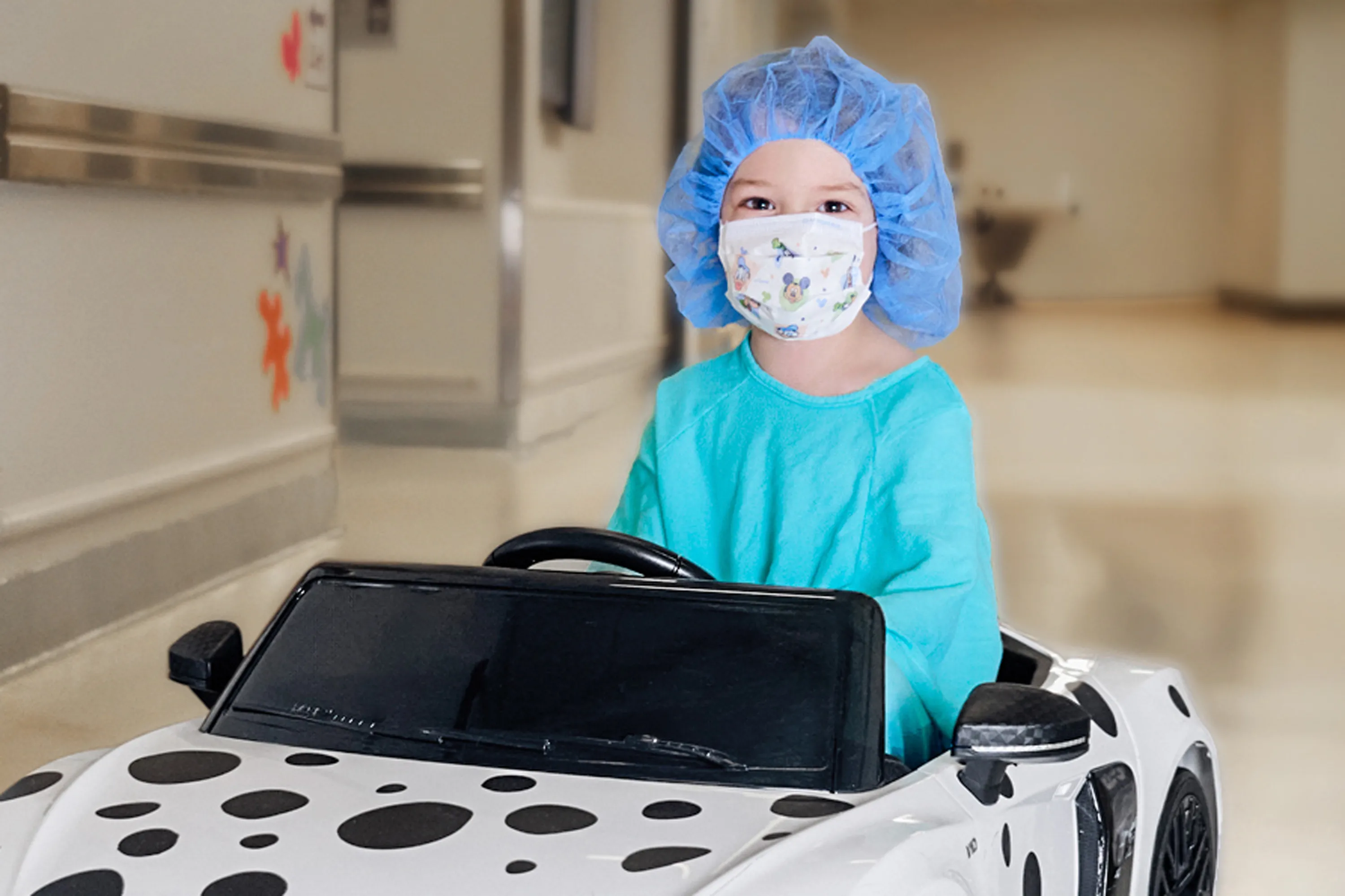 A child wearing a face mask and surgical cap sits in a toy car in a hospital hallway.