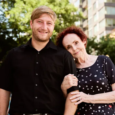 An older woman rests her head on a younger man’s shoulder as they both gently smile.