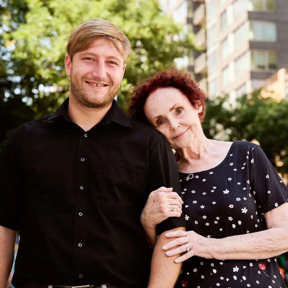 An older woman rests her head on a younger man’s shoulder as they both gently smile.