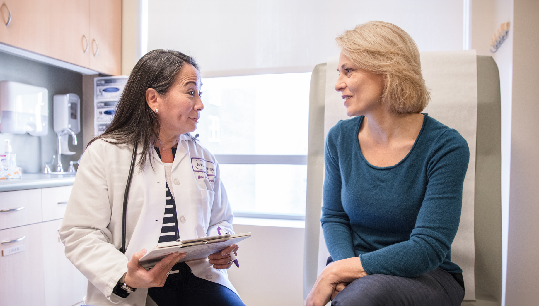 A doctor in a white lab coat and holding a clipboard talks to a woman sitting on an exam table.