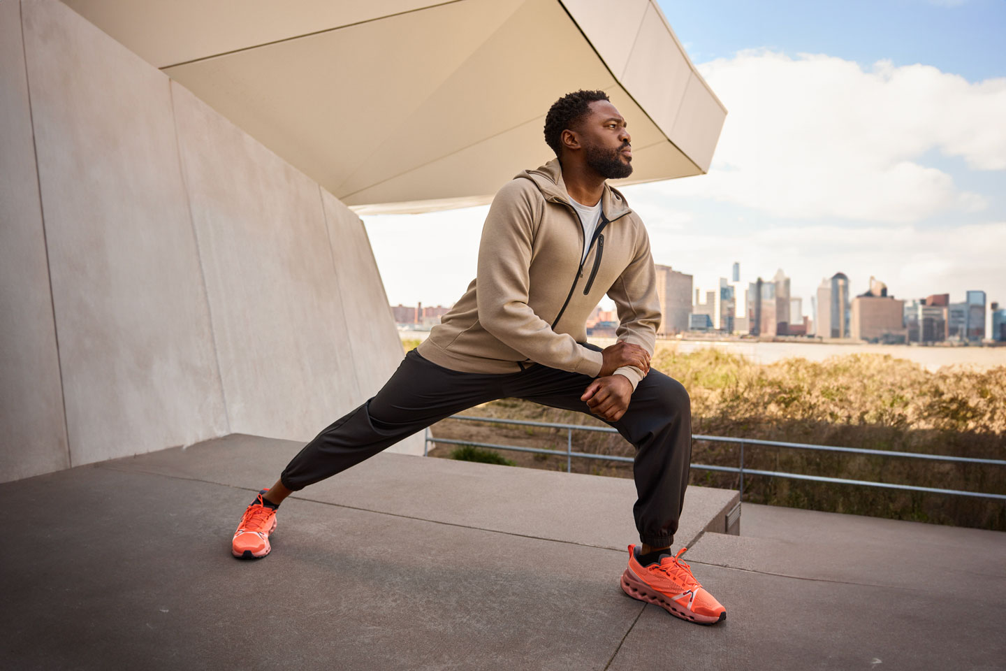 Dr. Kola Jegede wearing running attire, stretching in a forward lunge atop a concrete structure with city skyline behind him