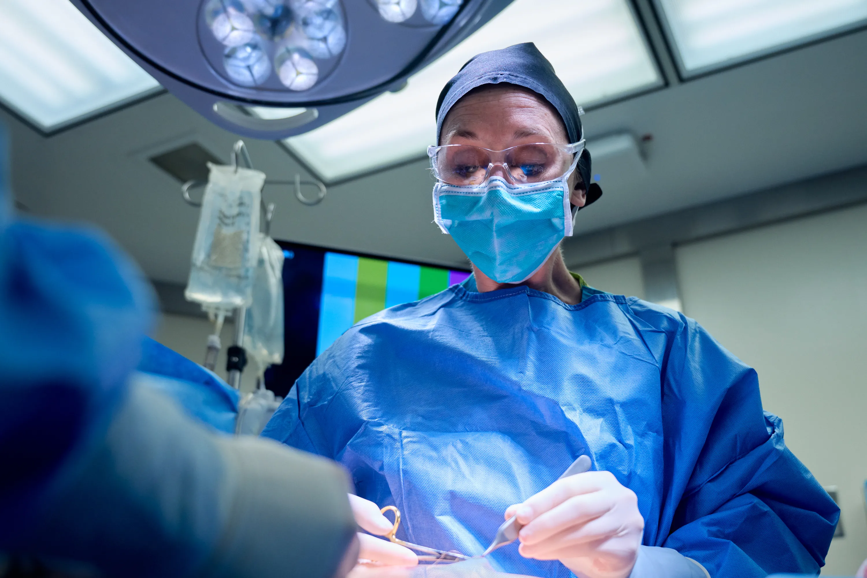 A doctor with a surgical mask, cap, and gown performs surgery in an operating room with monitor in the background.