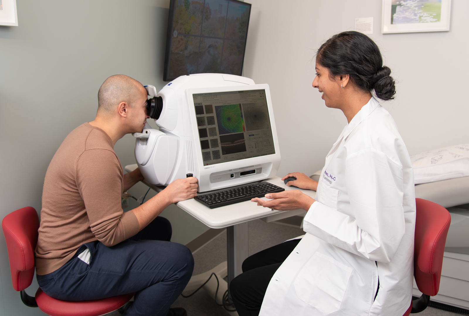 A patient sits at an eye exam machine with their face positioned against a viewer while physician assistant Swathi Bolneni looks at images on a screen next to the patient.