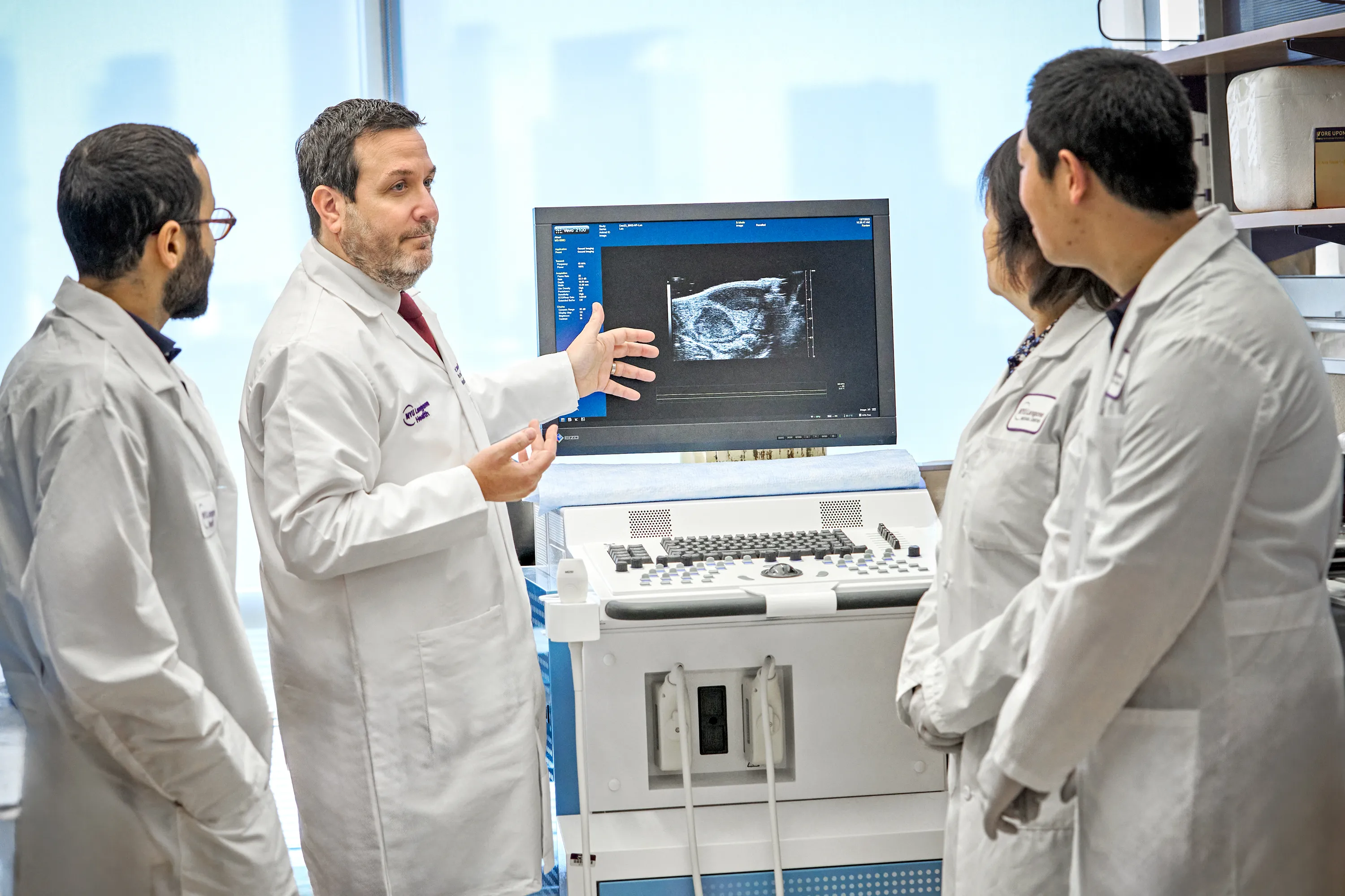 One doctor gestures toward a computer monitor displaying a radiology image, as three other doctors observe.