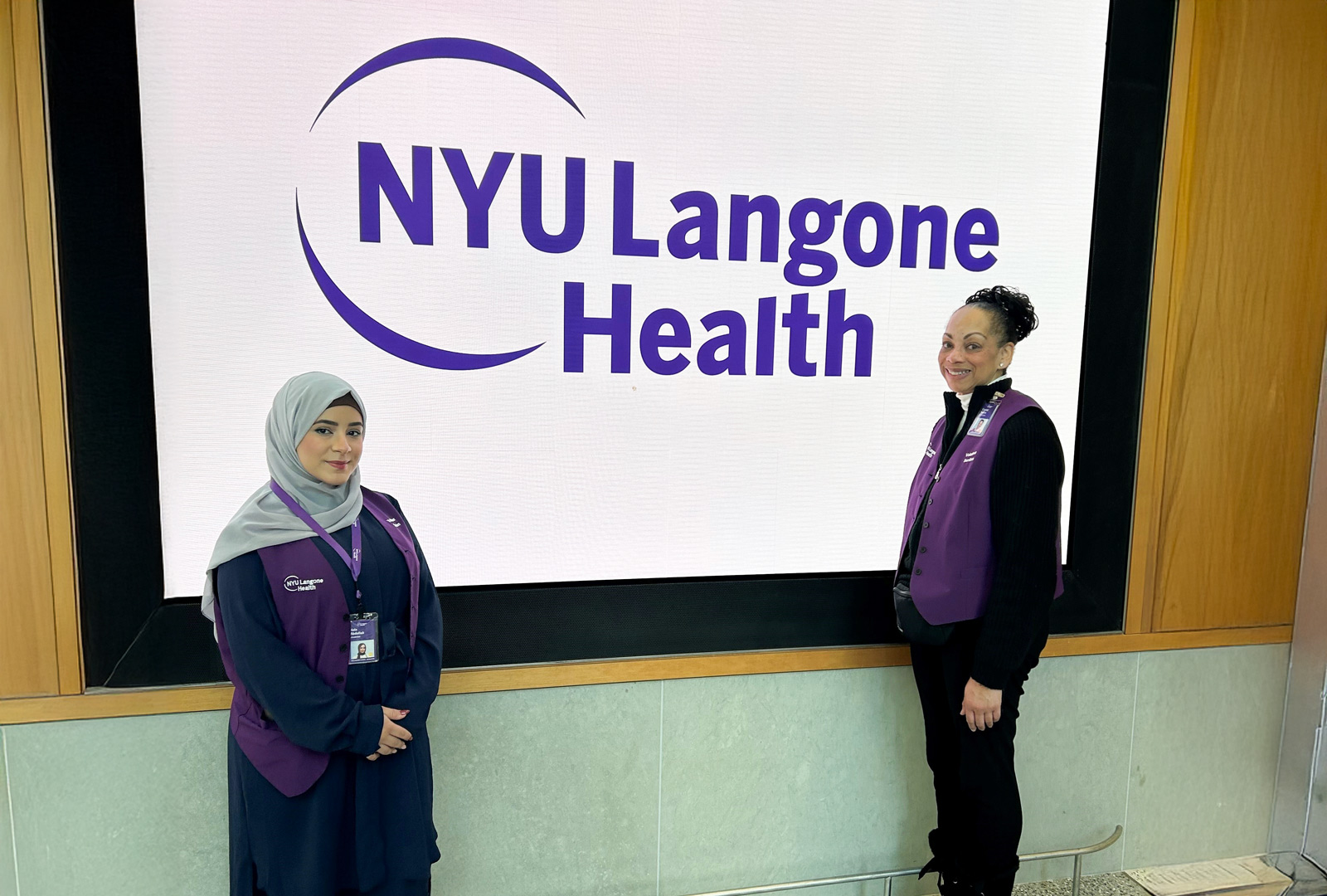 Two people wearing purple NYU Langone Health vests; one on the left wears a light gray headscarf and navy clothes, and the other on the right is wearing black, standing in front of an NYU Langone sign.