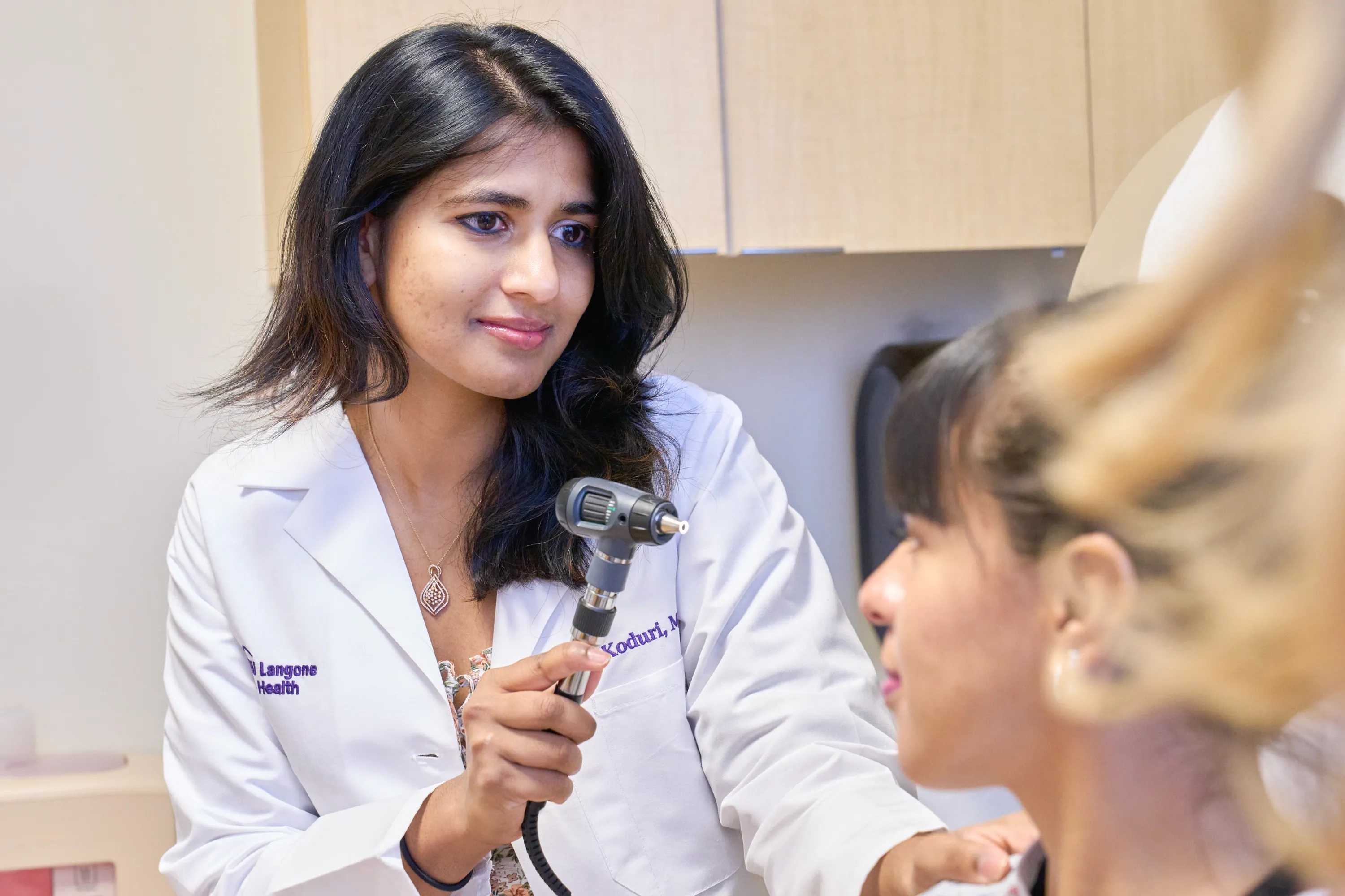 A doctor places her hand on a patient's shoulder while using a hand-held light to examine the patient’s eyes.