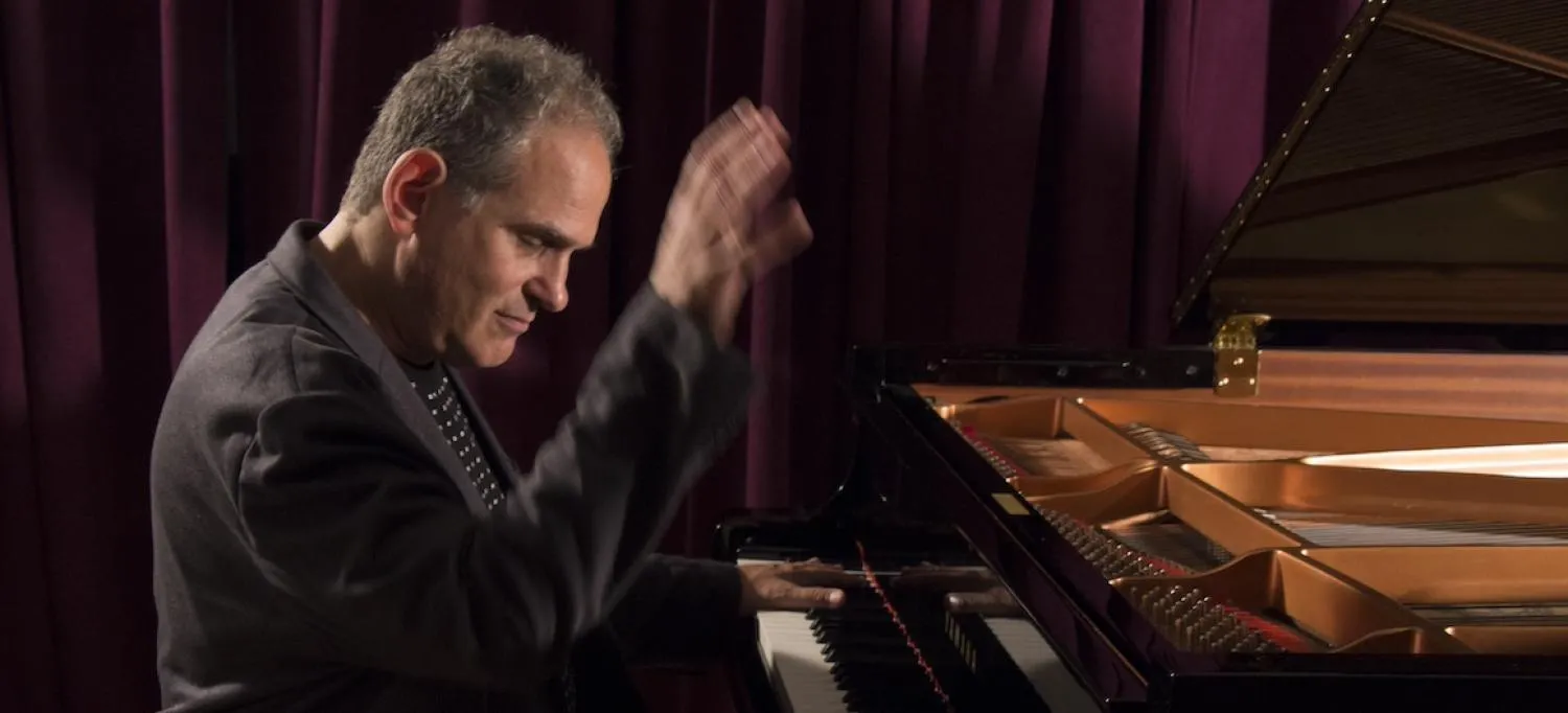 A man wearing a dark jacket is raising his right hand as he is playing a grand piano.