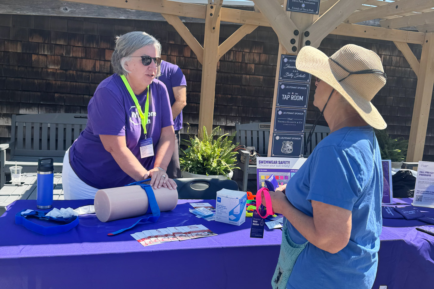 An NYU Langone provider applies a tourniquet on a training model at an outdoor community event while speaking with a woman.