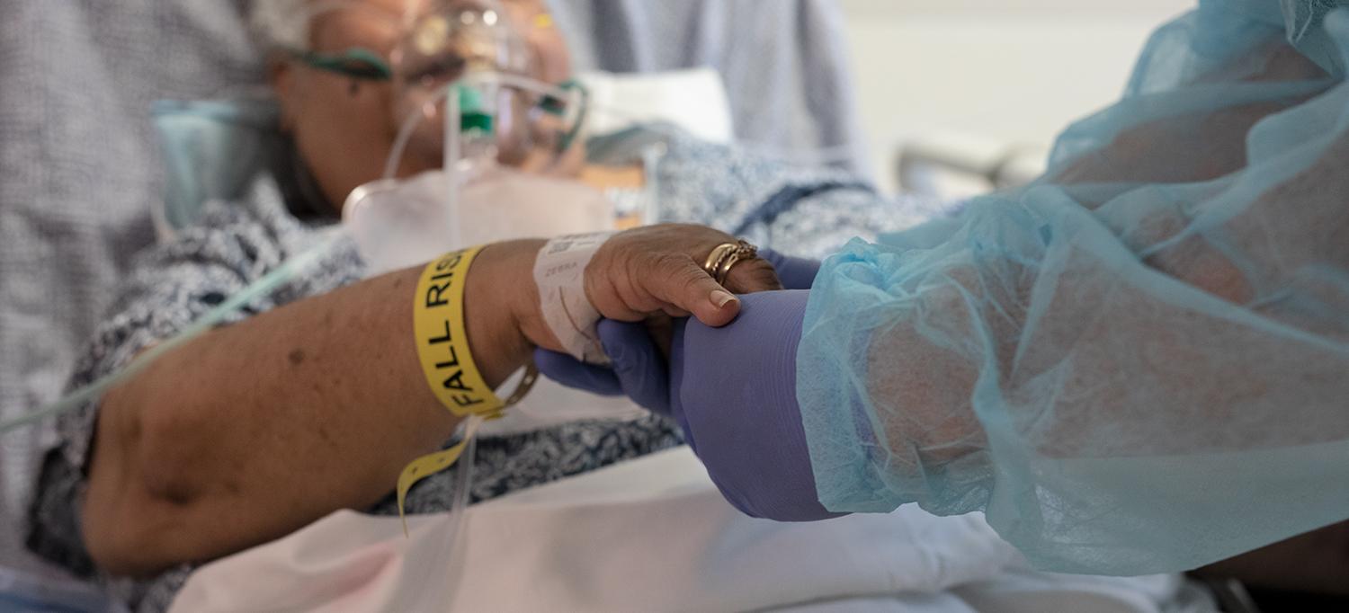 Patient Using Respirator in Hospital Bed Holds Nurse’s Gloved Hand