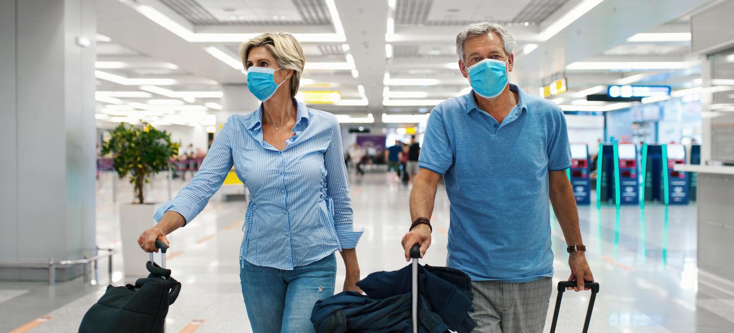 Two People Wearing Face Masks Rolling Luggage Through Airport Terminal