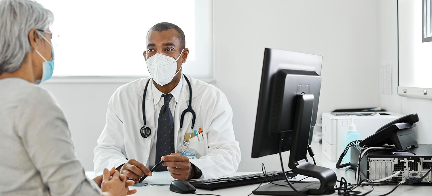 Doctor and Patient Wearing Face Masks, Sitting at Desk Together