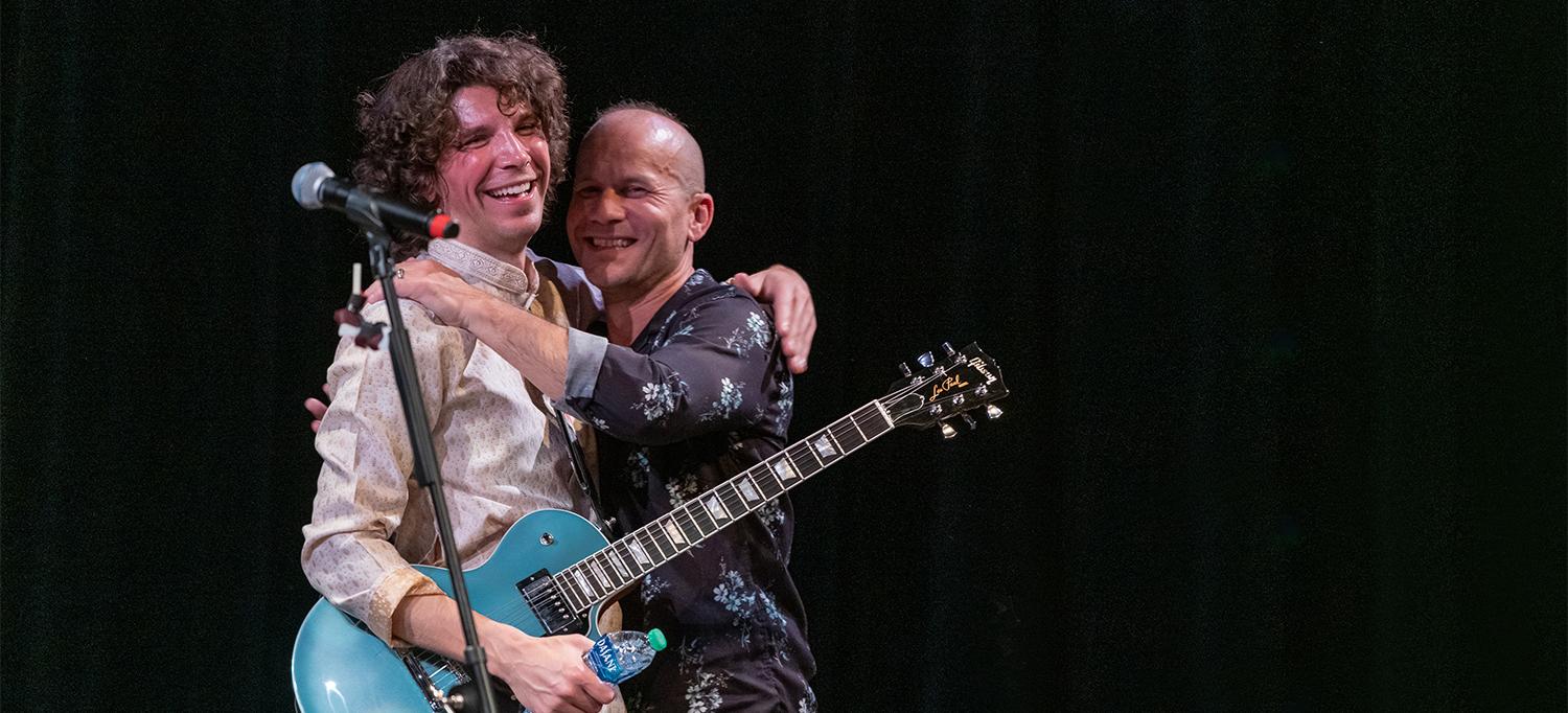 Jesse Kinch, Wearing Guitar on Strap, and Dr. Todd J. Carpenter Embrace on Stage During Performance