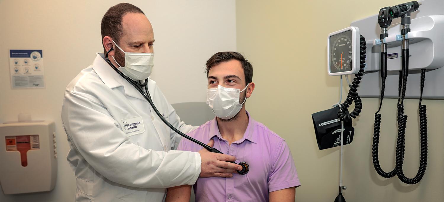 Dr. Dan G. Halpern Listening to Tyler Reynolds’s Heart with a Stethoscope in Exam Room, Both Are Wearing Face Masks