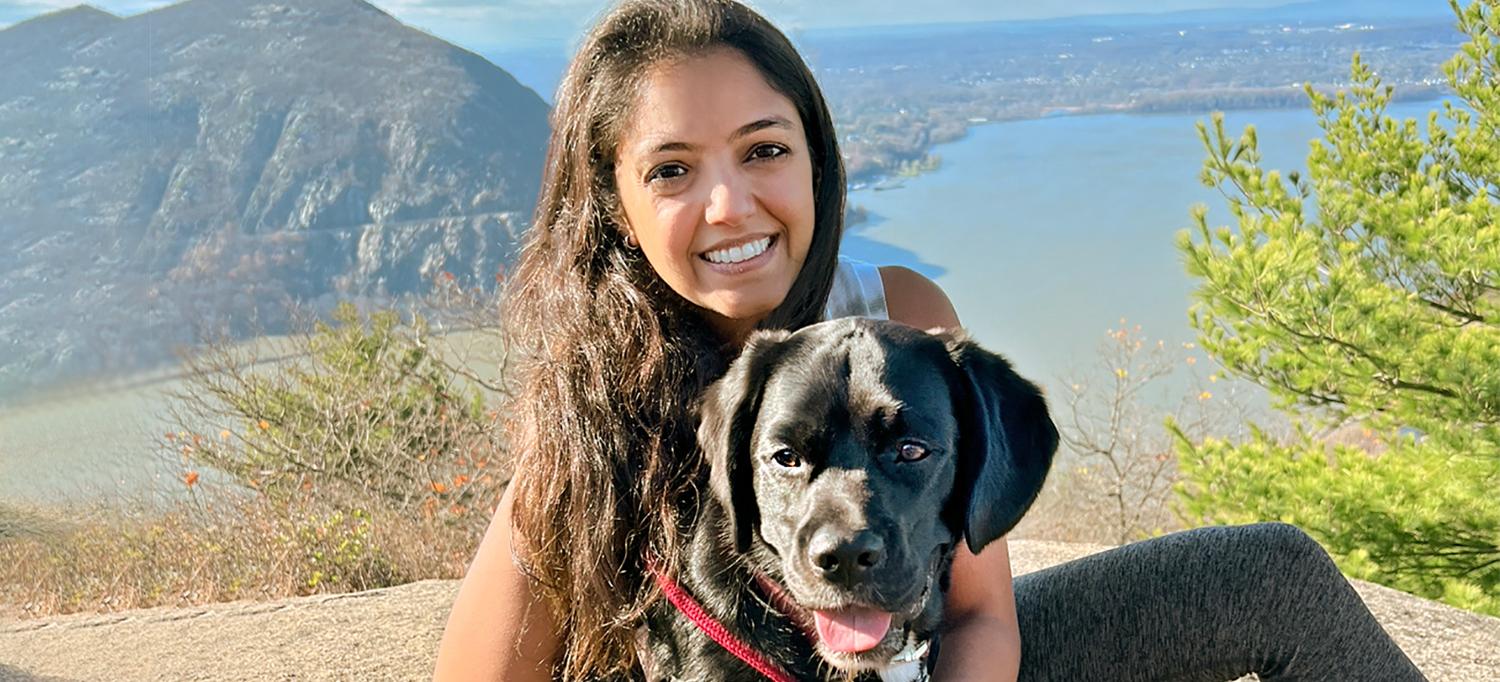 A Smiling Dr. Kinjan Parikh Poses with Her Dog Atop a Mountain Overlooking a River