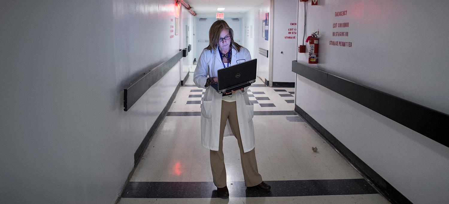 A Nurse Stands in the Hallway and Checks a Laptop
