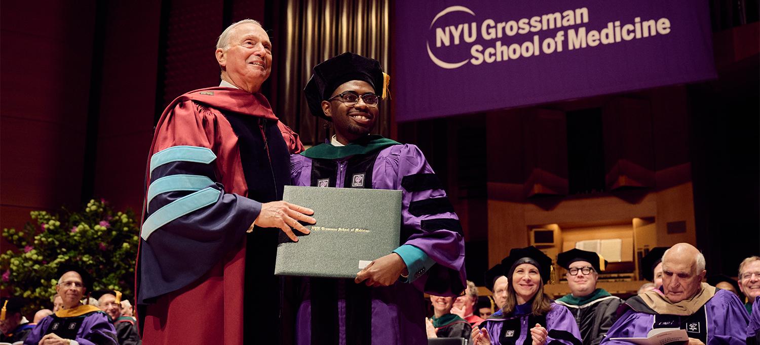 Dean Dr. Robert I. Grossman Poses with Graduate and Their Degree on Stage in Front of Colleagues