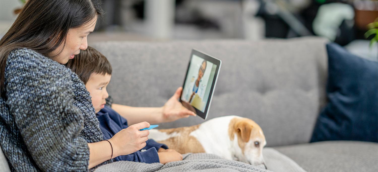 Mother Holding Thermometer and Son, Sitting on Couch with Dog and Using Tablet for Video Visit with Doctor