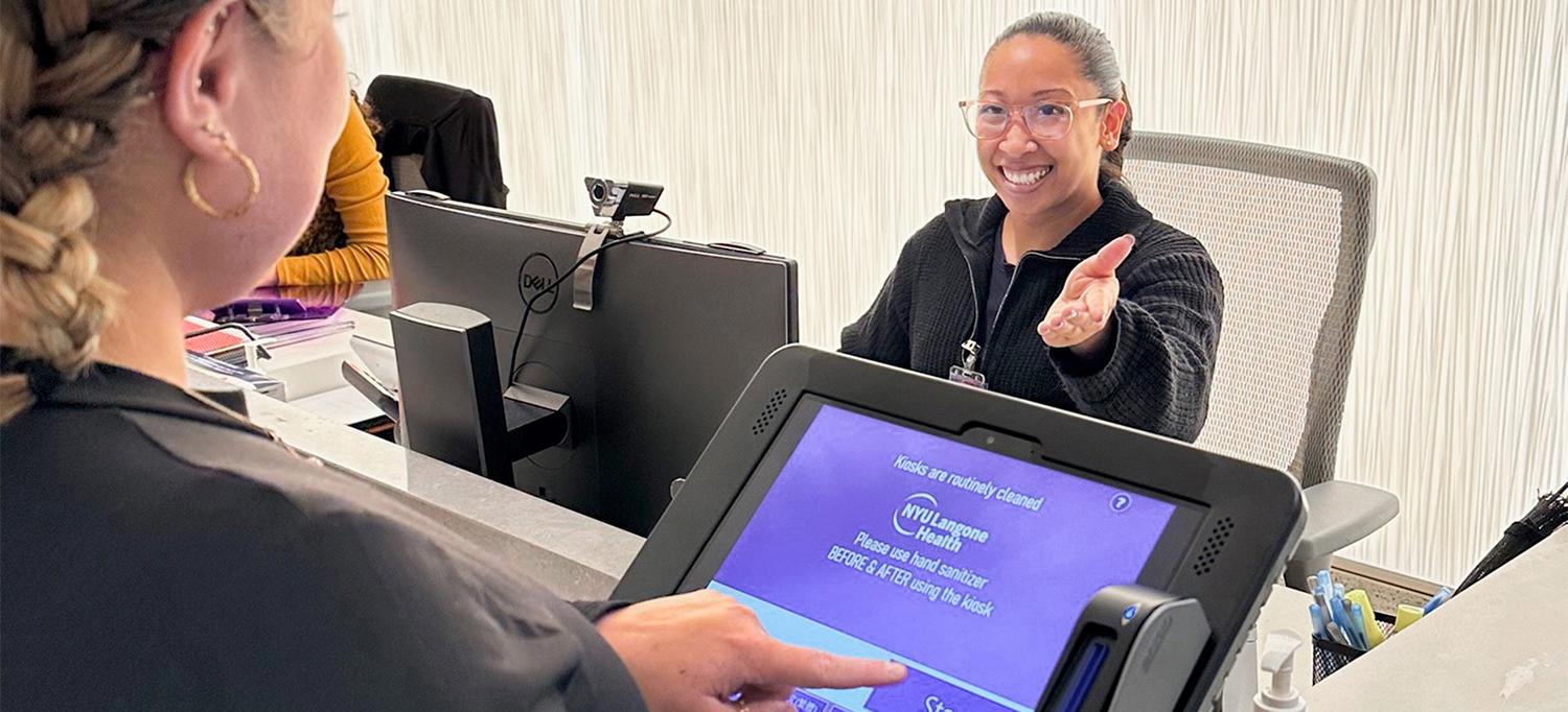 Nicole Davila is smiling and holding a hand out, gesturing to a patient to use the front desk kiosk to check in.