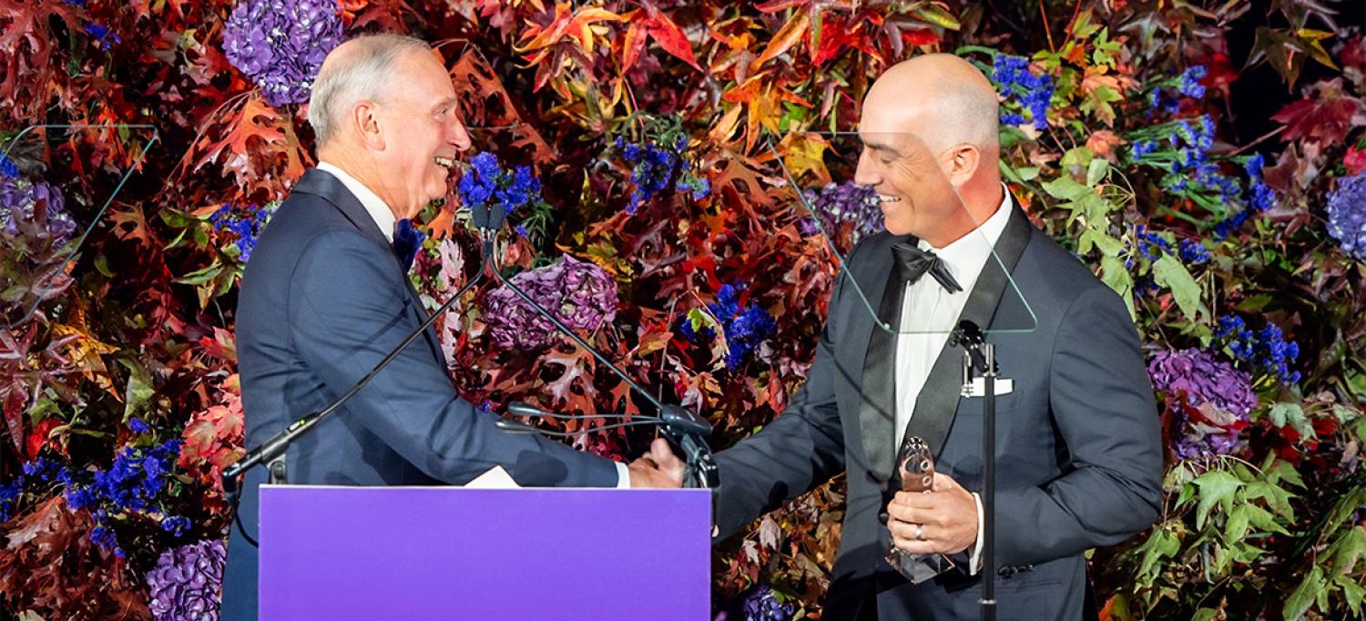 Dr. Grossman and Bezos smile and shake hands at a podium on stage in front of decorative foliage while Bezos holds the award