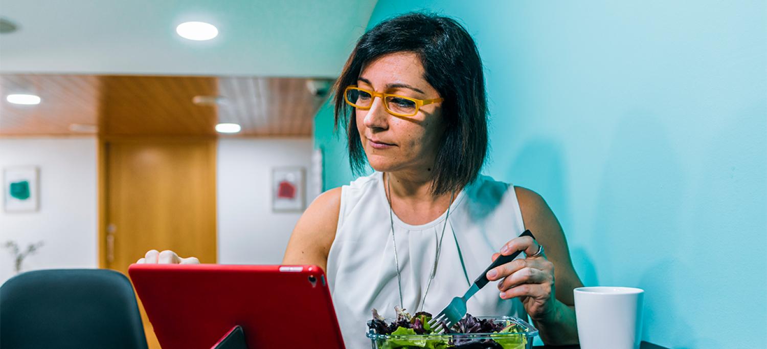 Woman sitting down and eating salad while using tablet device