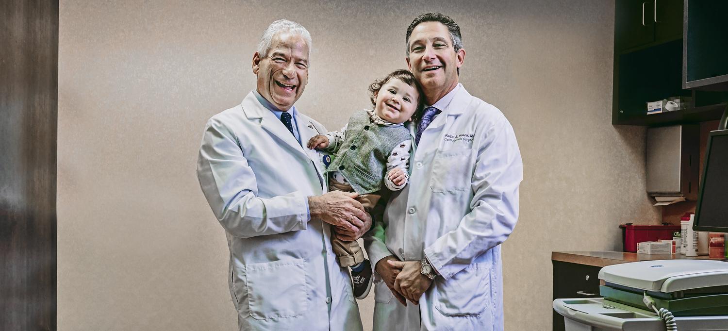 Sammie being held by Dr. Ludomirsky, who stands next to Dr. Mosca in a medical office.
