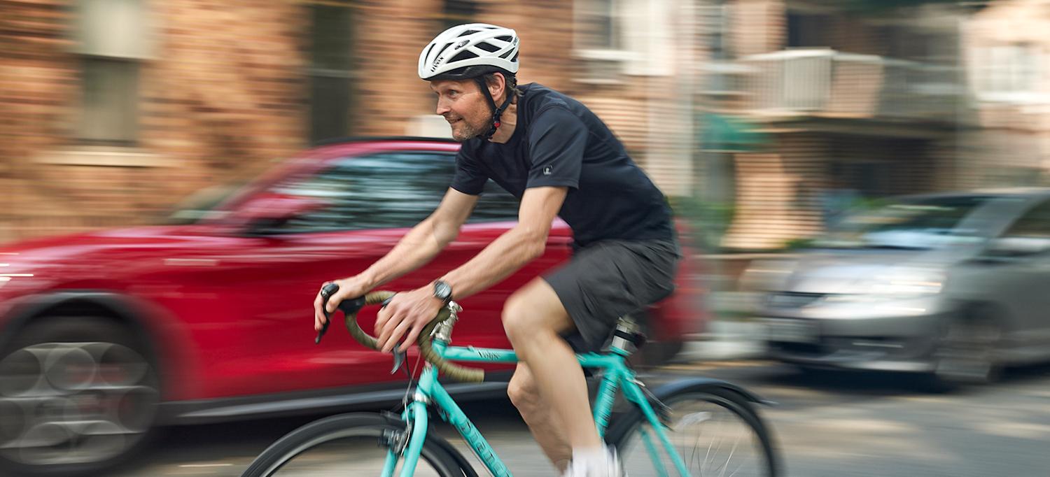 Keblish wearing a helmet and athletic apparel, riding his bike on a city street past a blurry car