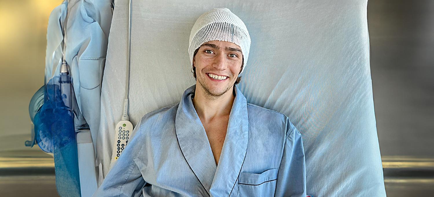 Barton Cowperthwaite, smiling and wearing a head covering and a hospital gown while reclining in a hospital bed