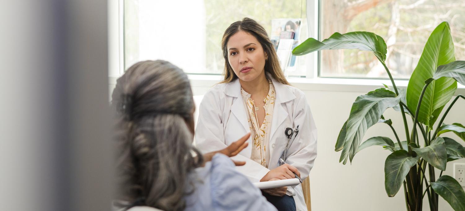 A healthcare provider in a white coat sits listening with a pad and paper to a patient whose back is to the camera.