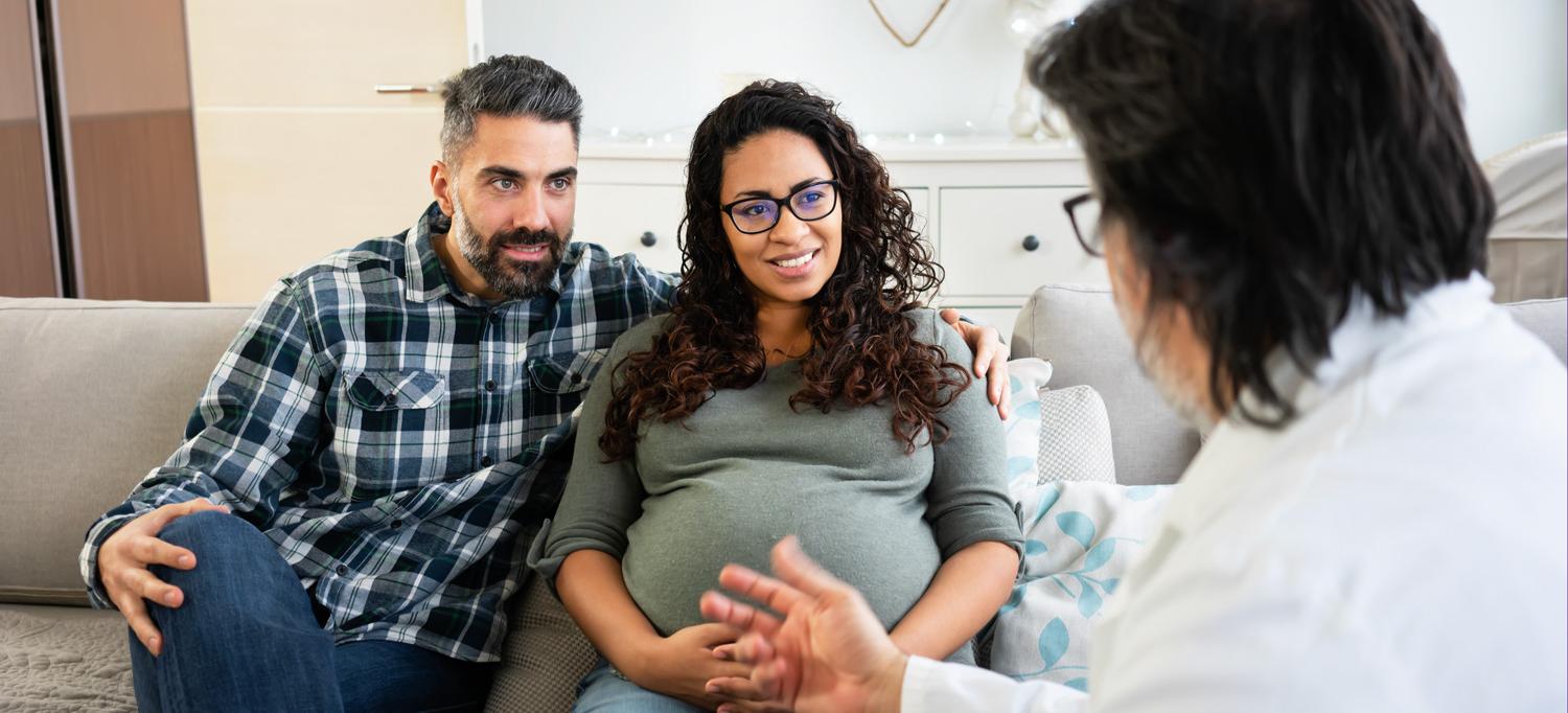 A pregnant and her partner speak with a doctor in their home.
