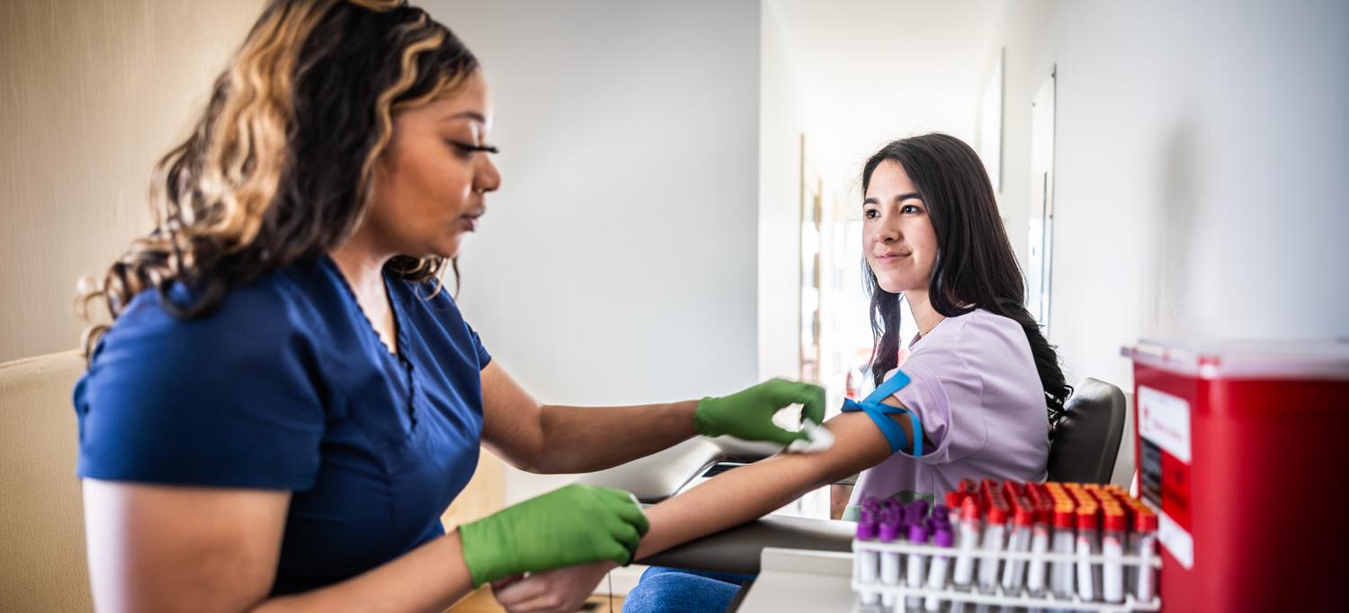 A phlebotomist prepares to draw blood from a young adult woman.