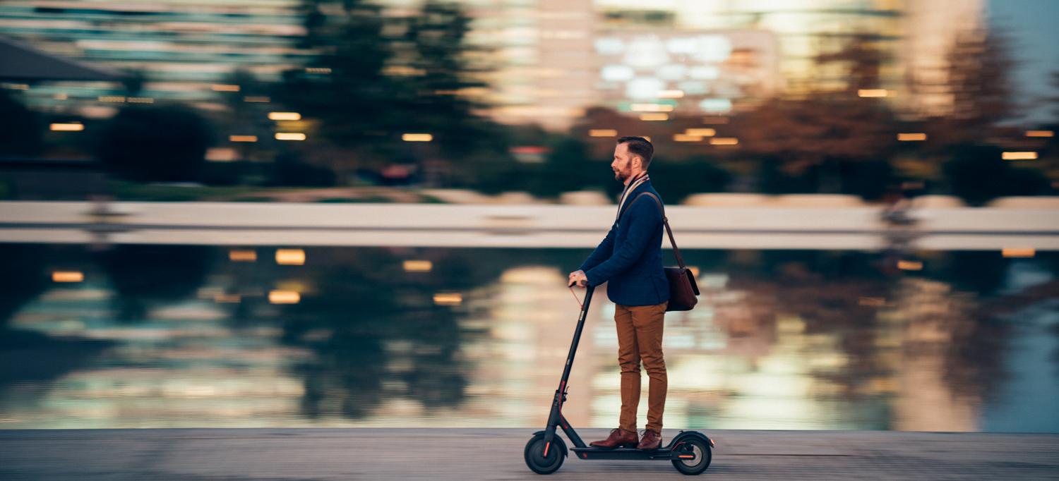 A man rides a scooter in the city with blurred lights behind him.