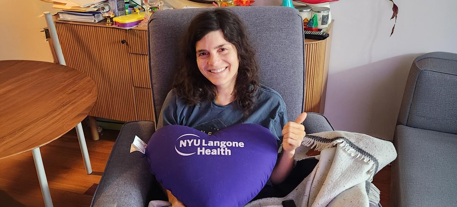 Hale sits in a chair holding a large purple pillow that says NYU Langone Health. 