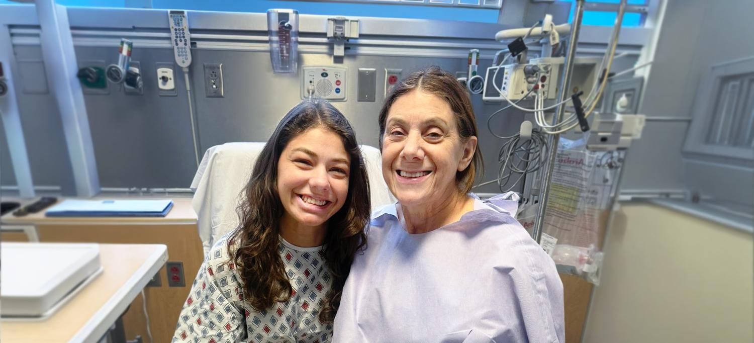 Both wearing hospital gowns, Velasquez and Schwicke sit on a hospital bed. 