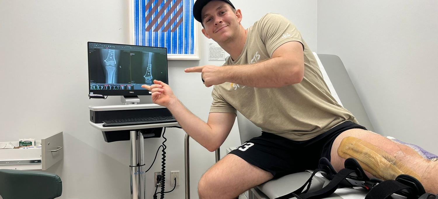 Nell sits on an exam chair pointing to a monitor with an image of his bones.