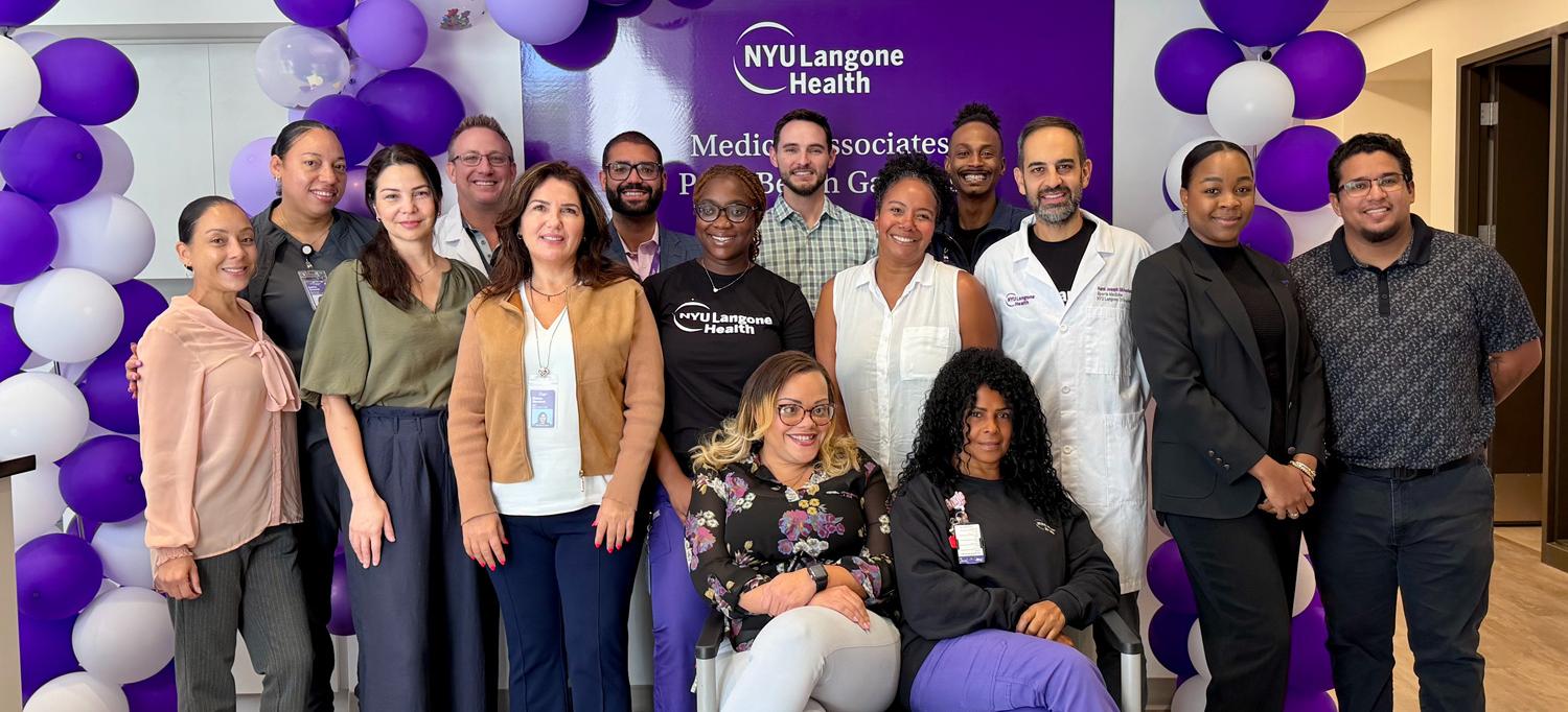 The staff at NYU Langone Medical Associates—Palm Beach Gardens stand smiling, in front of purple and white balloons and a sign with the name of the location. 