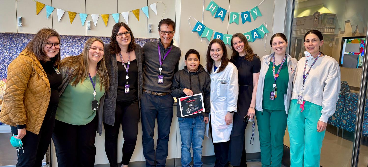 Darwin stands in a room surrounded by staff and well-wishers, with a sign reading “Happy Birthday” on the wall behind him. 