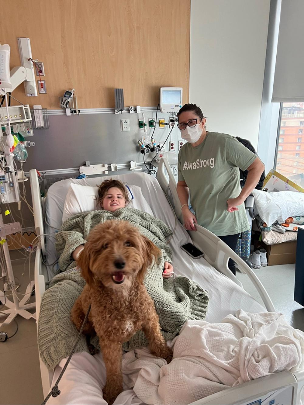 Ava in Hospital Bed with Therapy Dog and Mother Standing Beside Her