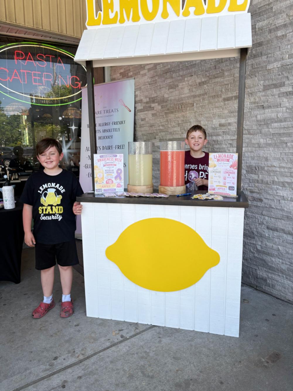 Logan and another boy are next to and behind a lemonade stand.
