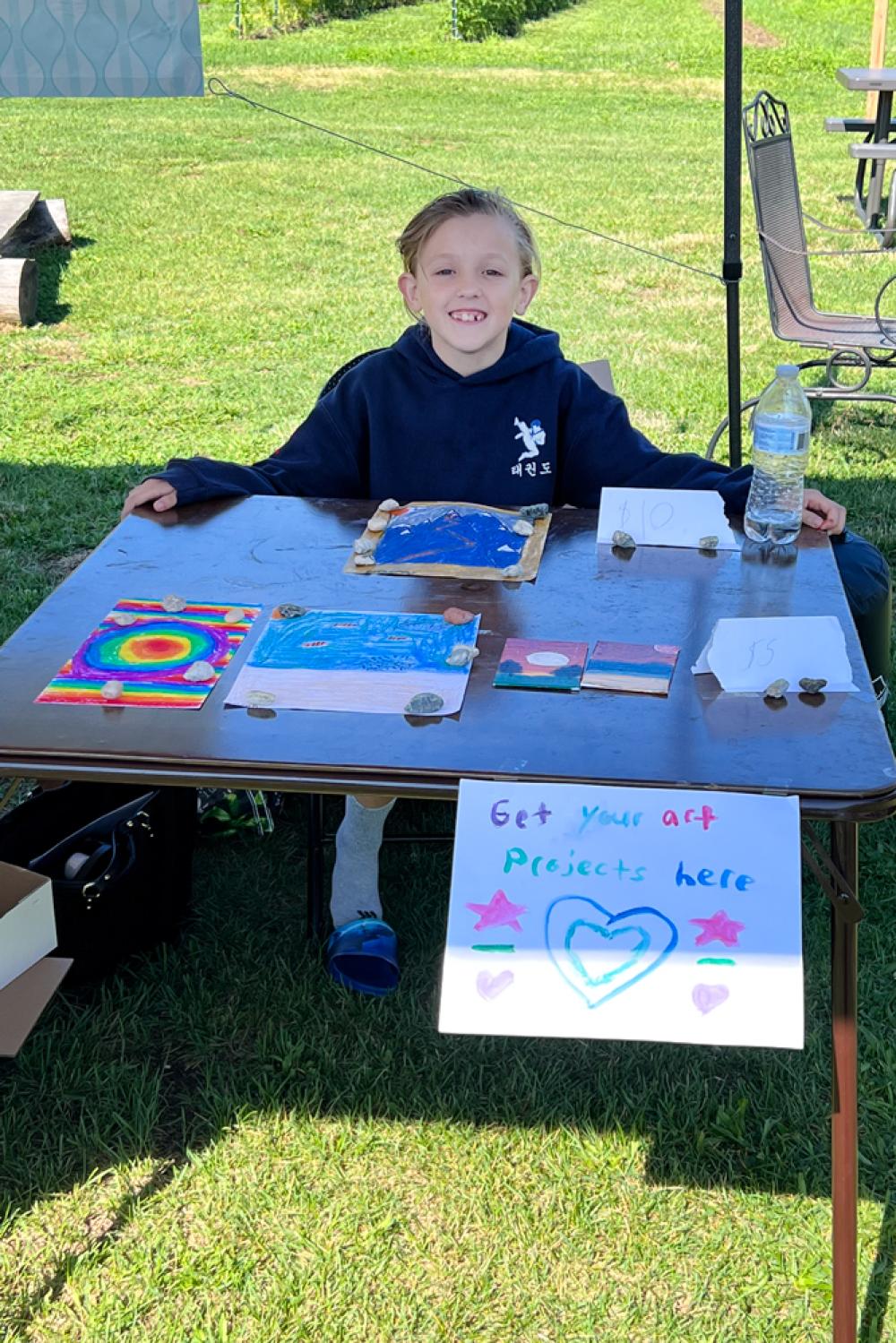 Logan sits outdoors behind a table with art projects on it. 