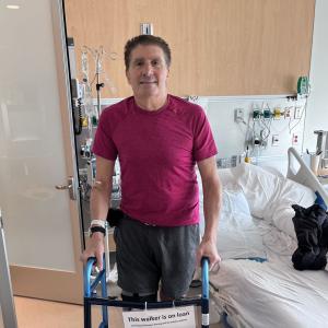 Using a walker, Richard Monti stands smiling in his hospital room.
