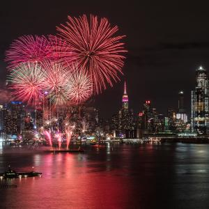 Red fireworks burst in the sky above the Hudson River, with the Empire State Building and midtown Manhattan in the background.