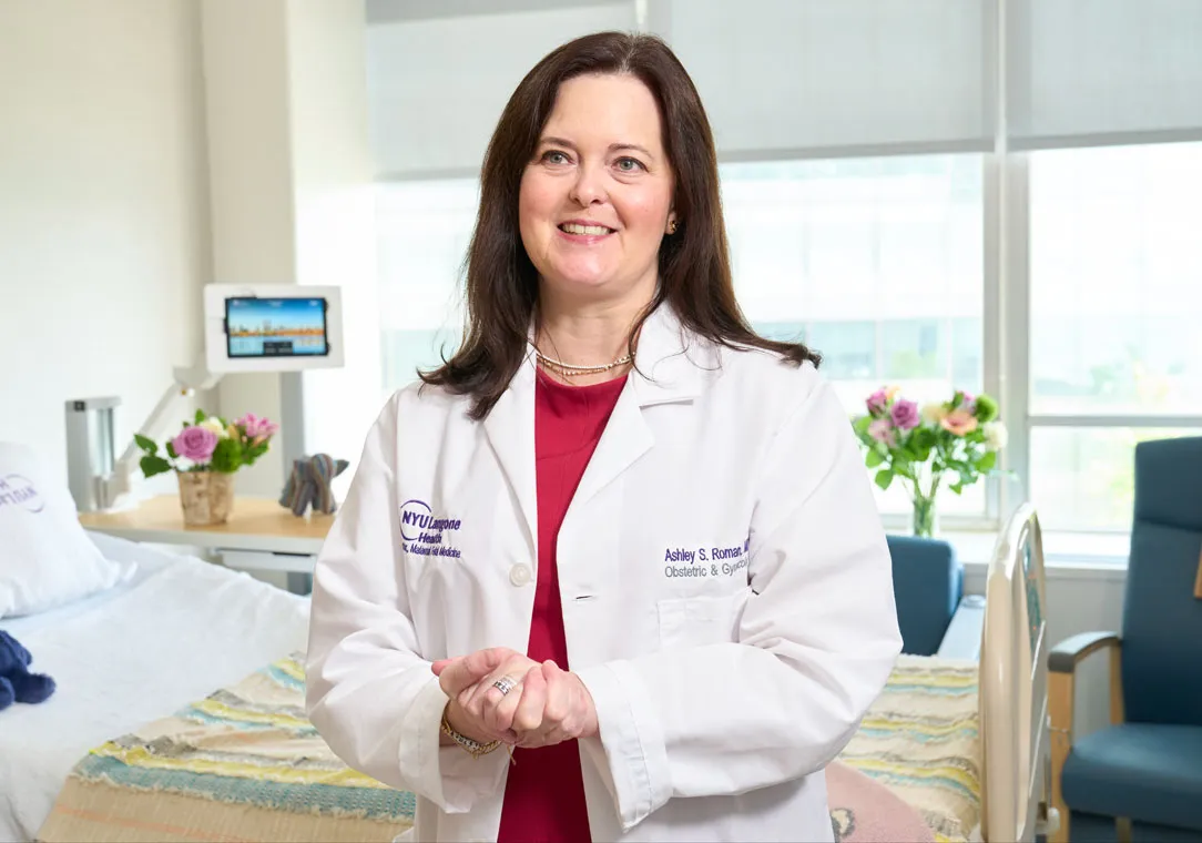 Dr. Ashley Roman stands with hands clasped in a hospital room with a bed, medical monitor, and large window.